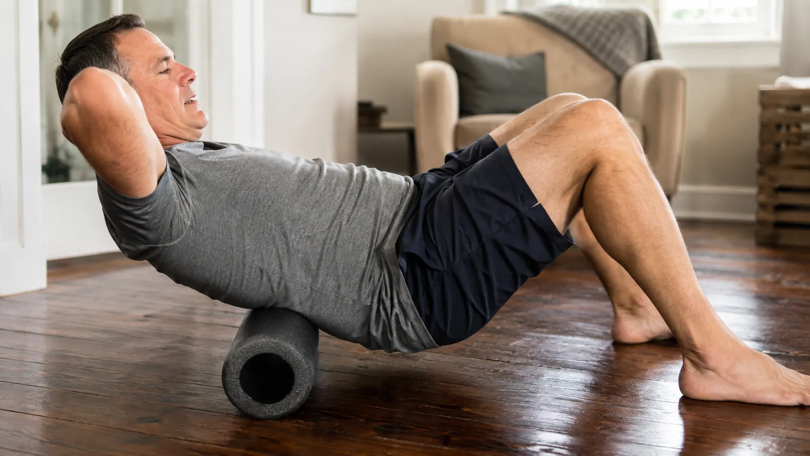 A man sitting on the floor of a home living room, leaning back onto a foam roller placed behind his lower back, soft morning light coming through a window